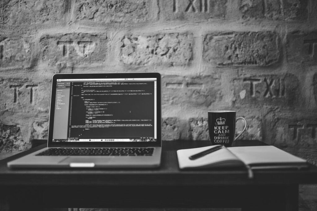 "Laptop displaying code on a desk with a 'Keep Calm and Drink Coffee' mug and notebook, set against a brick wall, representing a coding workspace environment."