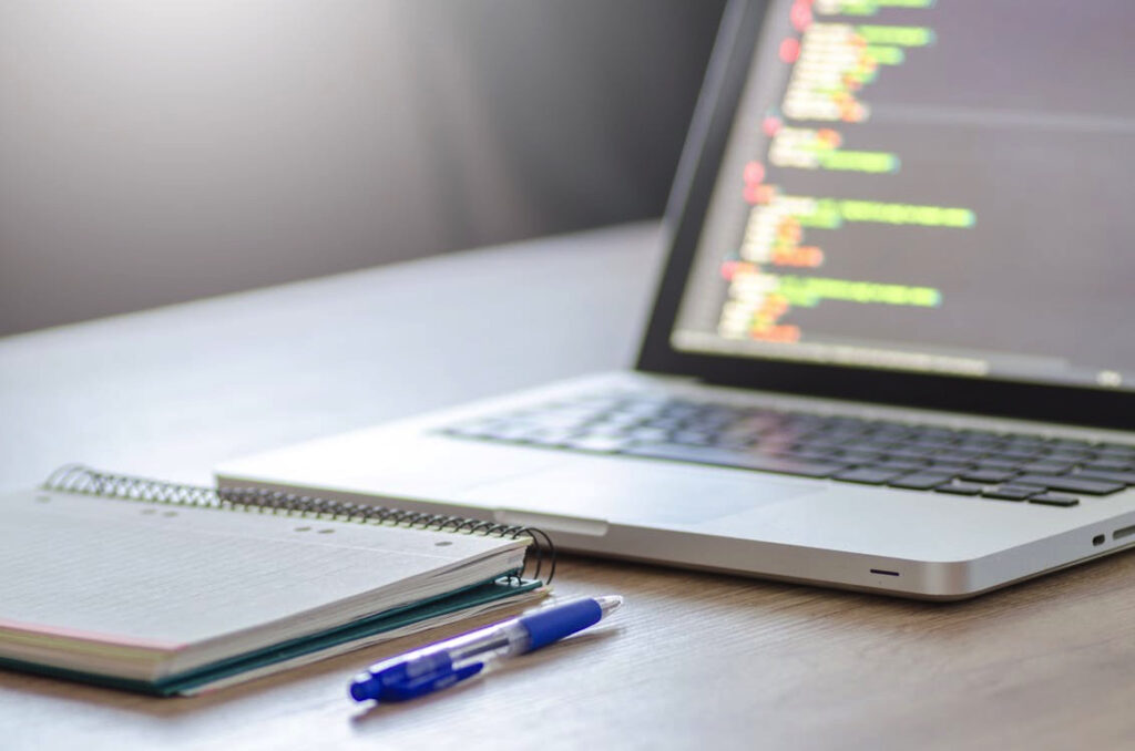 Laptop displaying code next to a notebook and pen on a wooden desk, ideal for programming and study sessions.