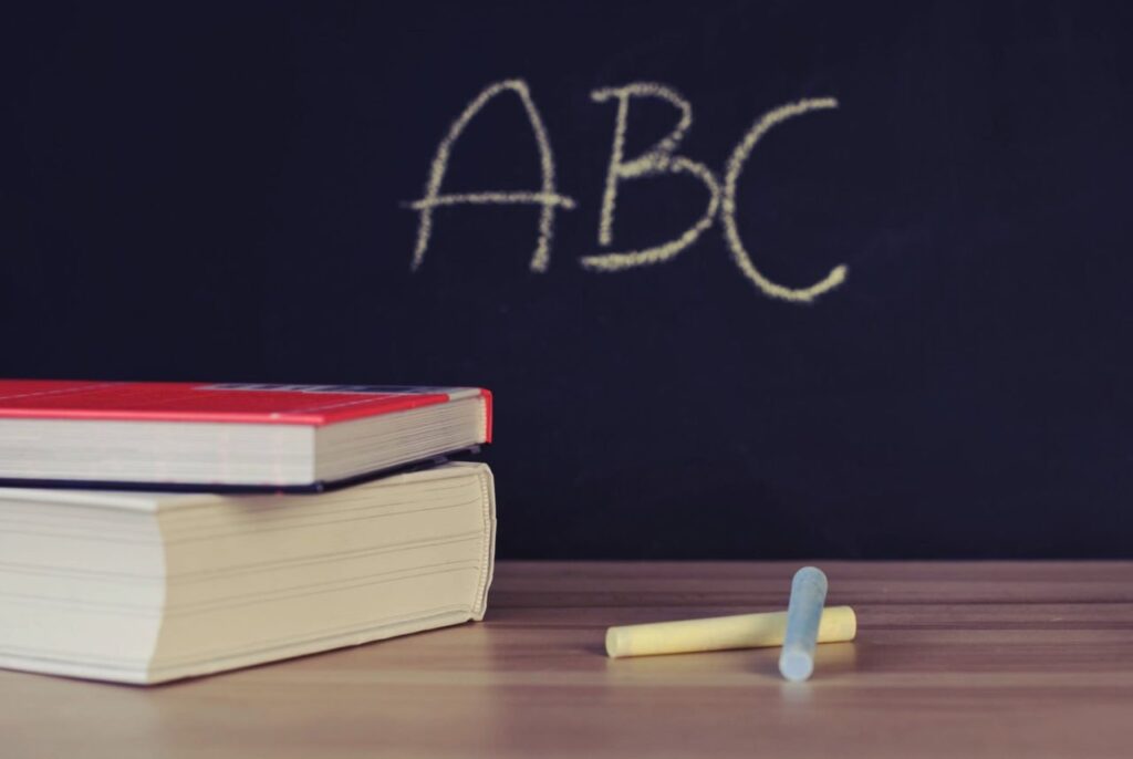 "Stack of books and chalk pieces on a wooden desk in front of a blackboard with 'ABC' written in yellow chalk, symbolizing education and learning concepts."