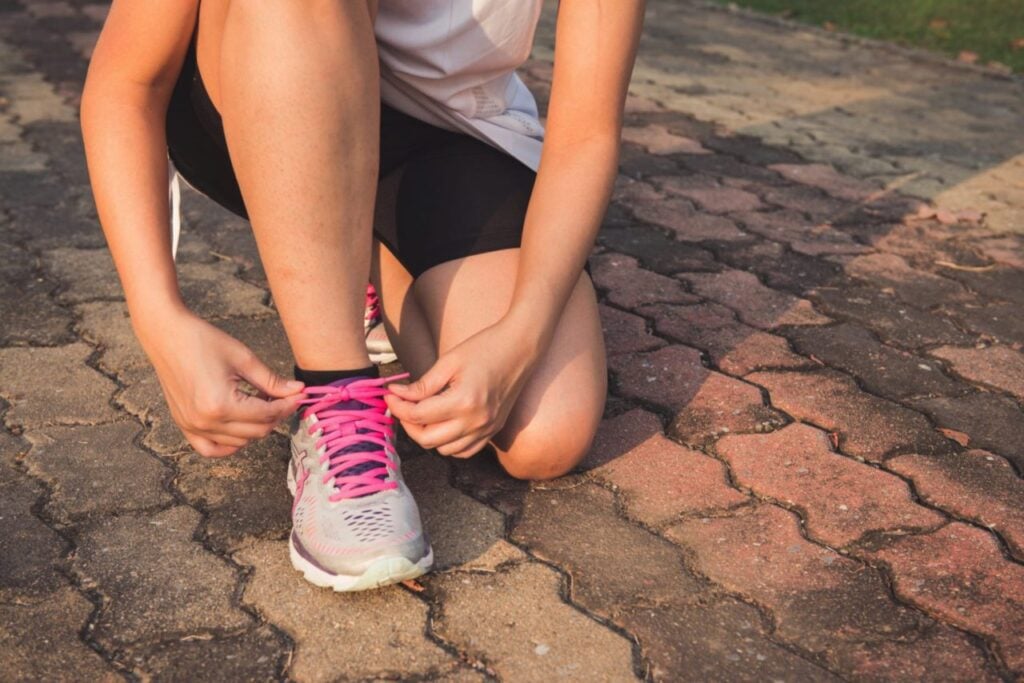 "Person tying pink shoelaces on running shoes while kneeling on a brick path outdoors, preparing for exercise or jogging."