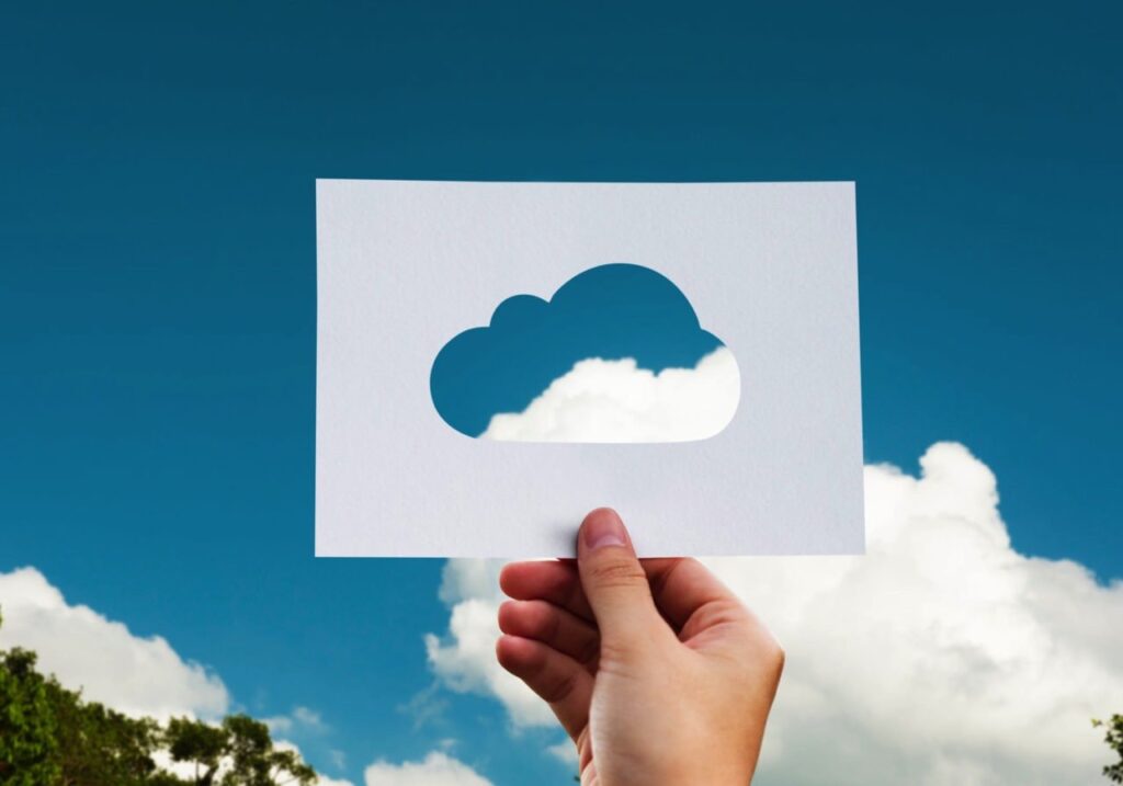 "Hand holding a paper with a cloud shape cutout against a blue sky background, symbolizing cloud computing and technology integration."