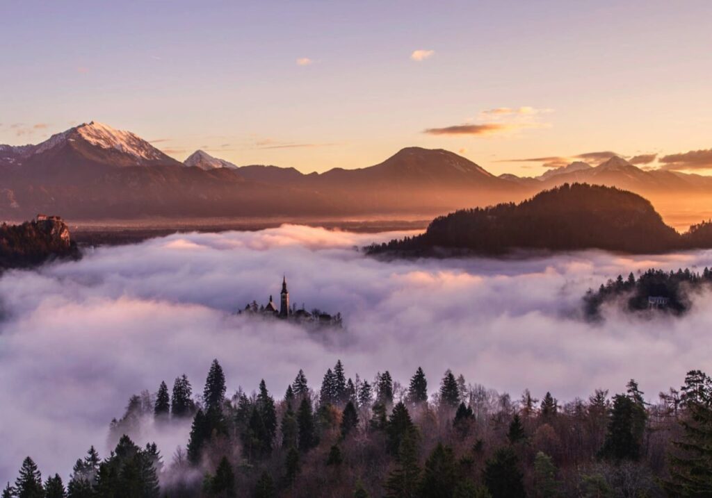 "Scenic view of a church emerging through fog surrounded by mountains and forest at sunrise, showcasing tranquil natural beauty and landscape serenity."