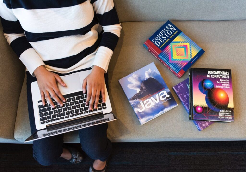 Person typing on a laptop with programming books nearby, including "Compiler Design," "Java," and "Fundamentals of Computing II," on a couch.