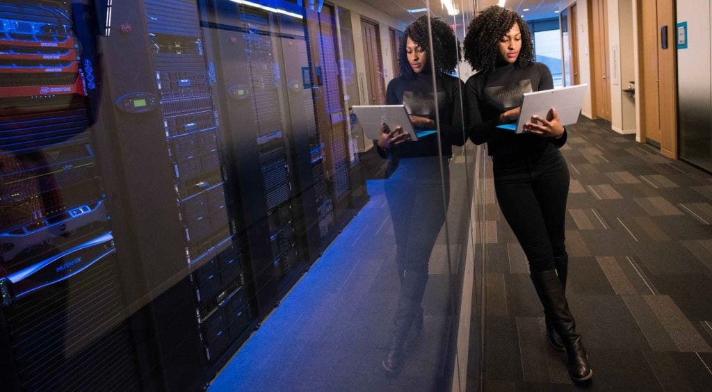 IT professional analyzing data on a tablet in a modern server room, with reflections on glass panels and rows of servers in the background.