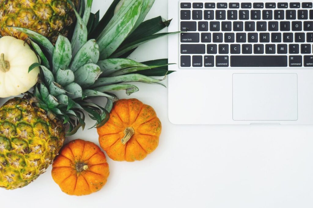 "Top view of laptop next to fresh pineapples and small pumpkins on a white background, symbolizing healthy lifestyle and remote work balance."