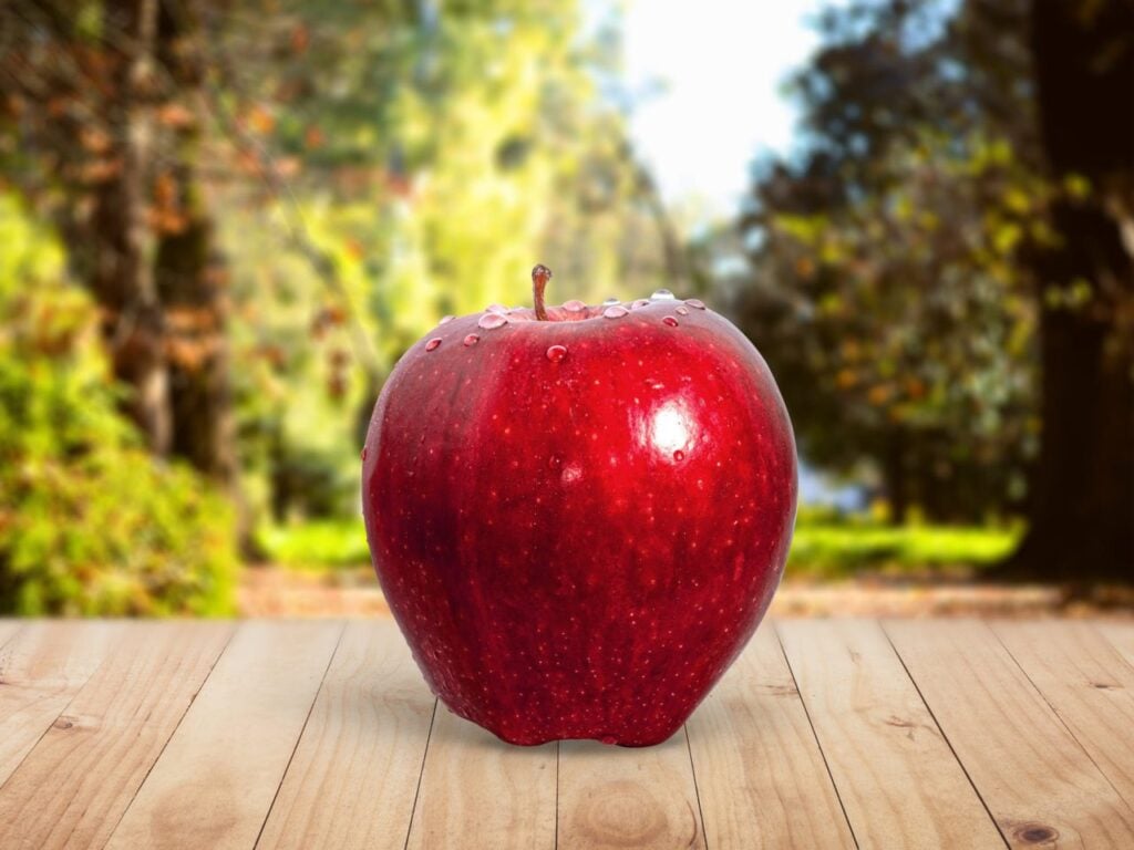 "Fresh red apple with water droplets on a wooden surface in a sunny outdoor setting, ideal for healthy eating and organic produce themes."