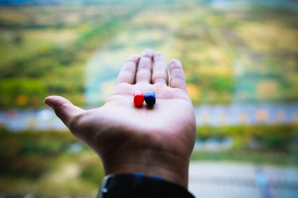 A close-up image of a hand holding red and blue capsules with a blurred outdoor background effect.